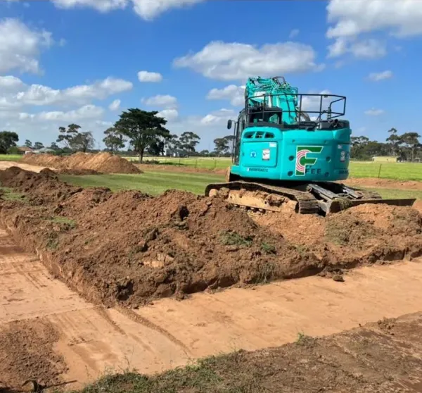 A green excavator is digging in a grassy field under a partly cloudy sky.