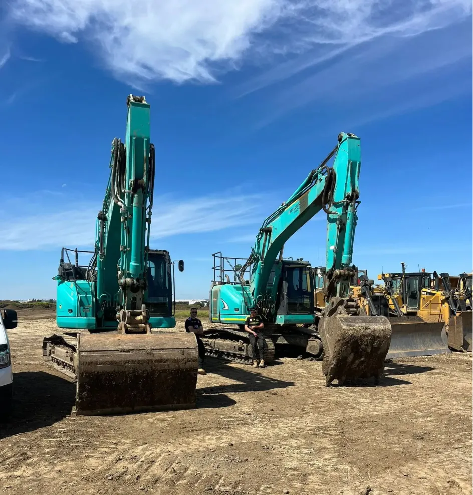 Two large teal excavators parked on a dirt construction site under a clear blue sky.
