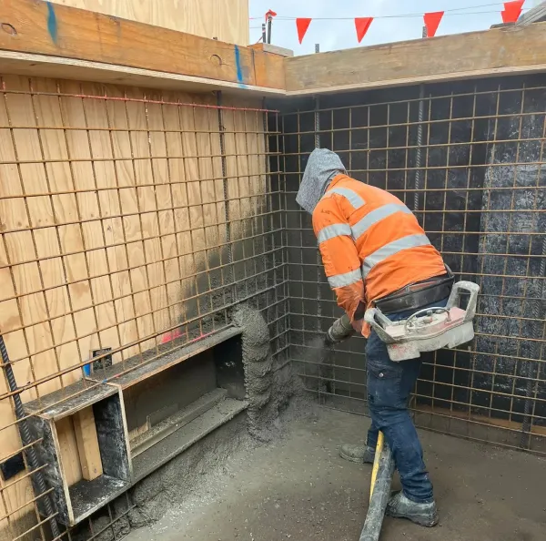 A construction worker sprays concrete on a reinforced wall inside a building under construction.